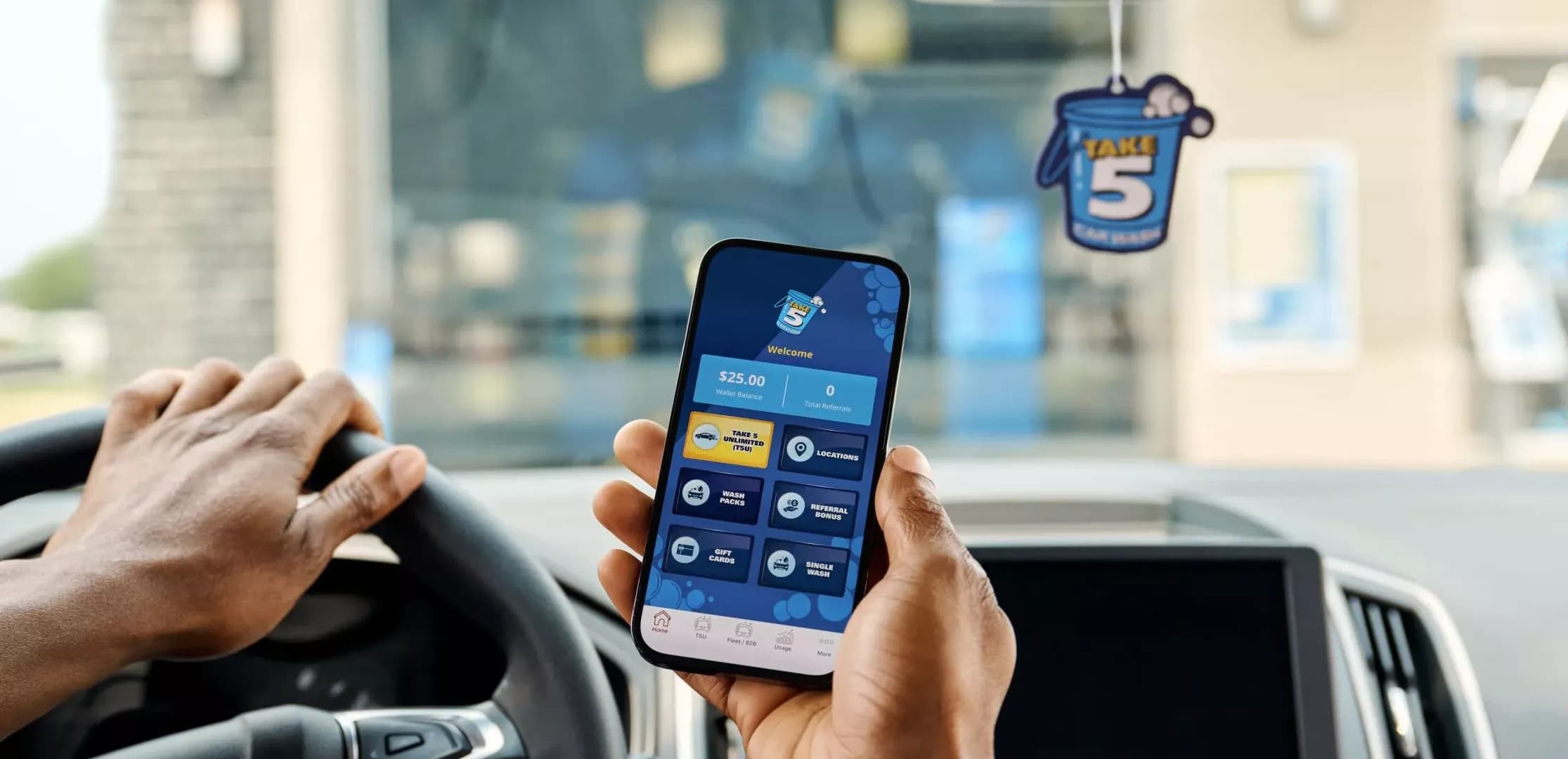 Person holding a phone with the Take 5 Car Wash app, inside a car with a Take 5 air freshener.